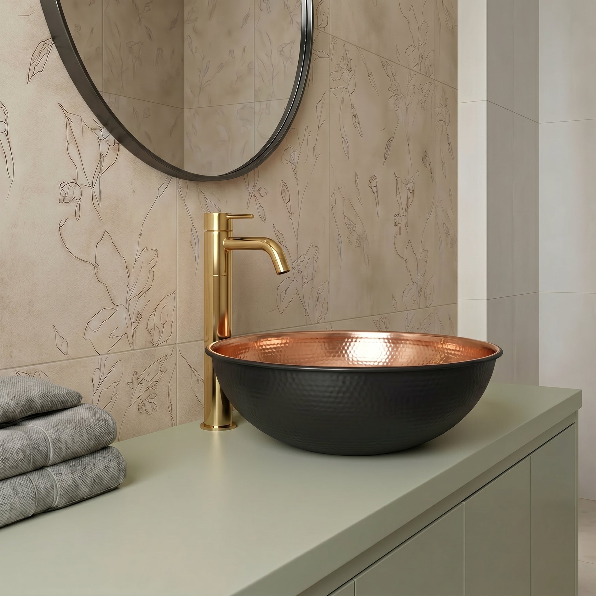 A modern bathroom vanity setup featuring a hammered black ceramic vessel sink with a hammered copper interior, paired with a brushed gold faucet. The sink is mounted on a sage green countertop against a beige floral-patterned tiled wall with a round mirror.